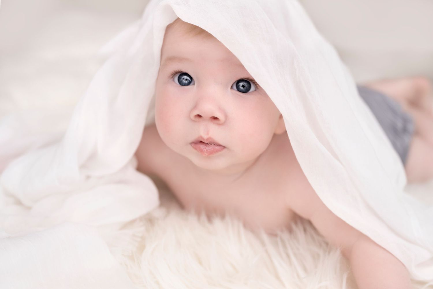 Adorable baby with blue eyes peeking under a soft white blanket on a fluffy white fur surface.