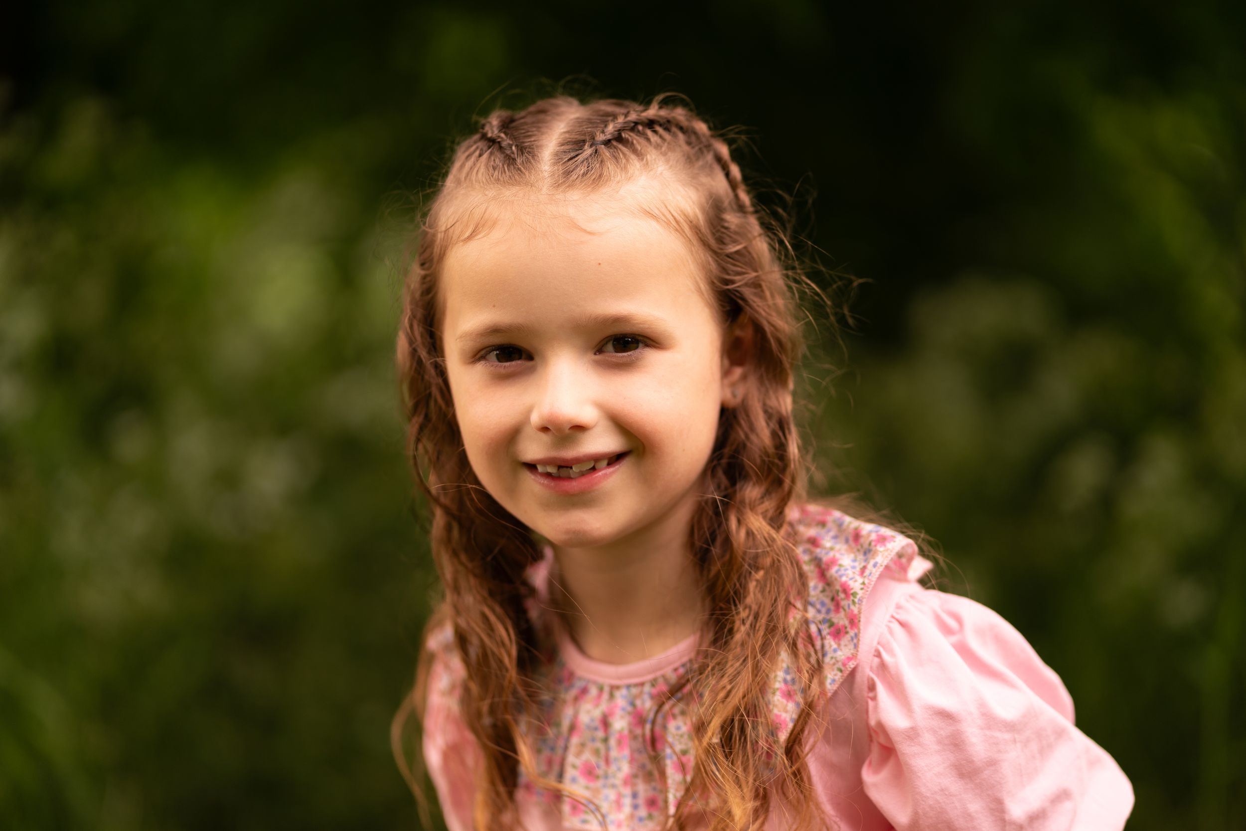 A smiling young girl with braided hair wearing a pink dress, photographed outdoors with green foliage background.