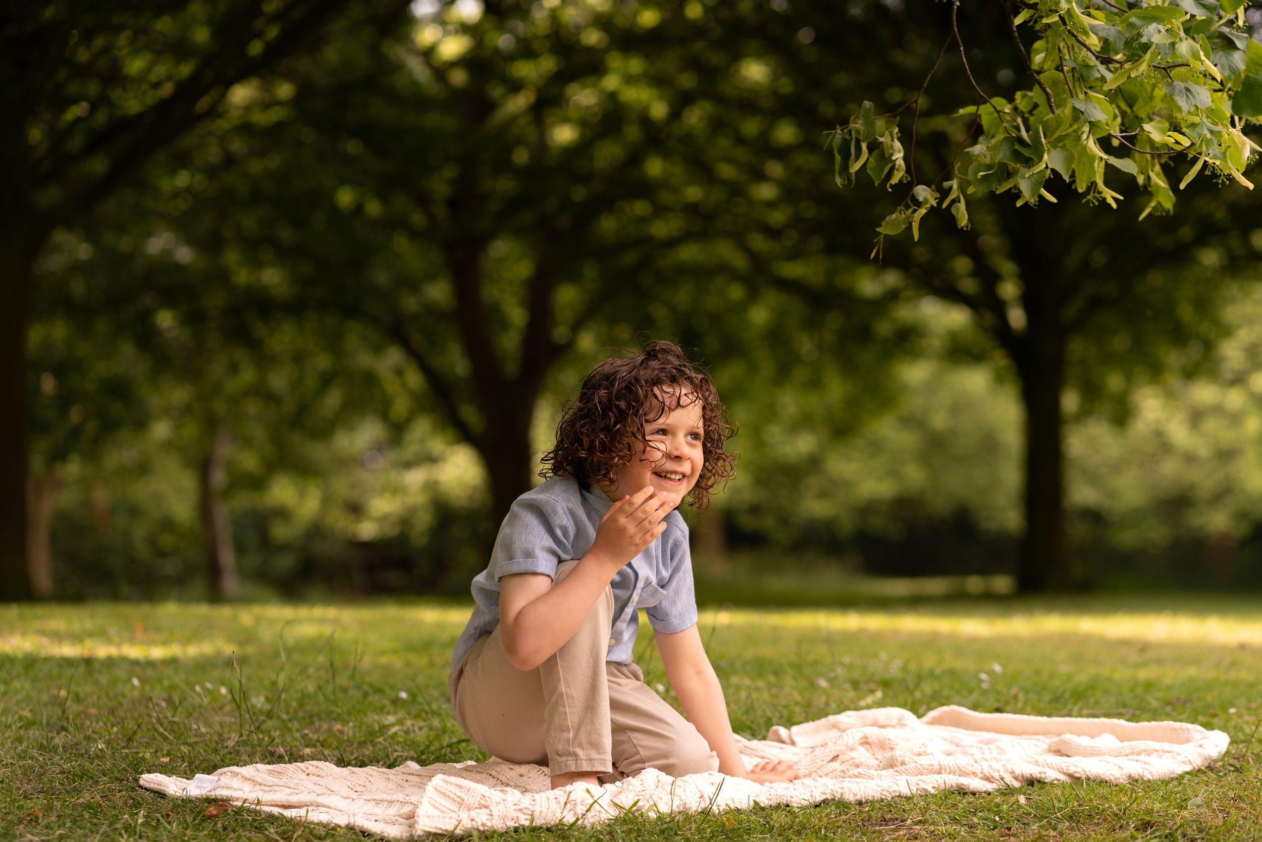 Woman with curly hair sitting on a blanket in a sunny green park, smiling and enjoying a relaxing outdoor picnic.