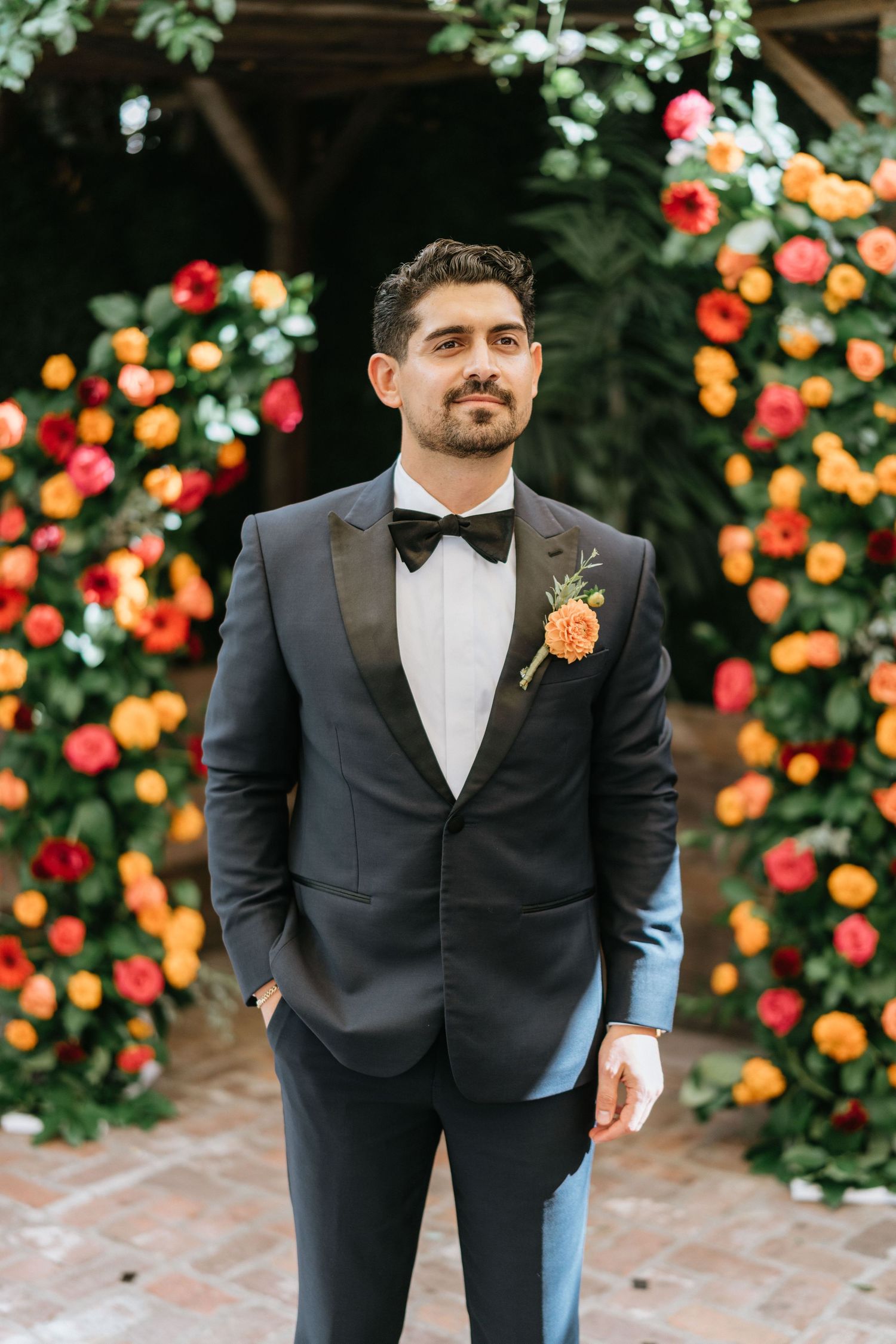 Full-length portrait of groom Sina standing in front of the colorful floral ceremony backdrop.