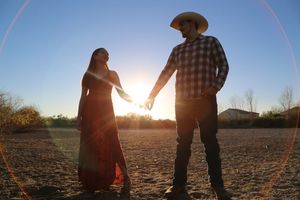 Couple in a cowboy hat and flowing red dress walk hand in hand across open desert land at golden hour.