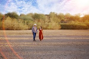 Man in a cowboy hat and plaid shirt embraces a woman in a rust red dress during an outdoor engagement shoot.