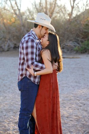 Couple embraces forehead to forehead outdoors, he in a cowboy hat and plaid shirt, she in a long rust red dress at sunset.