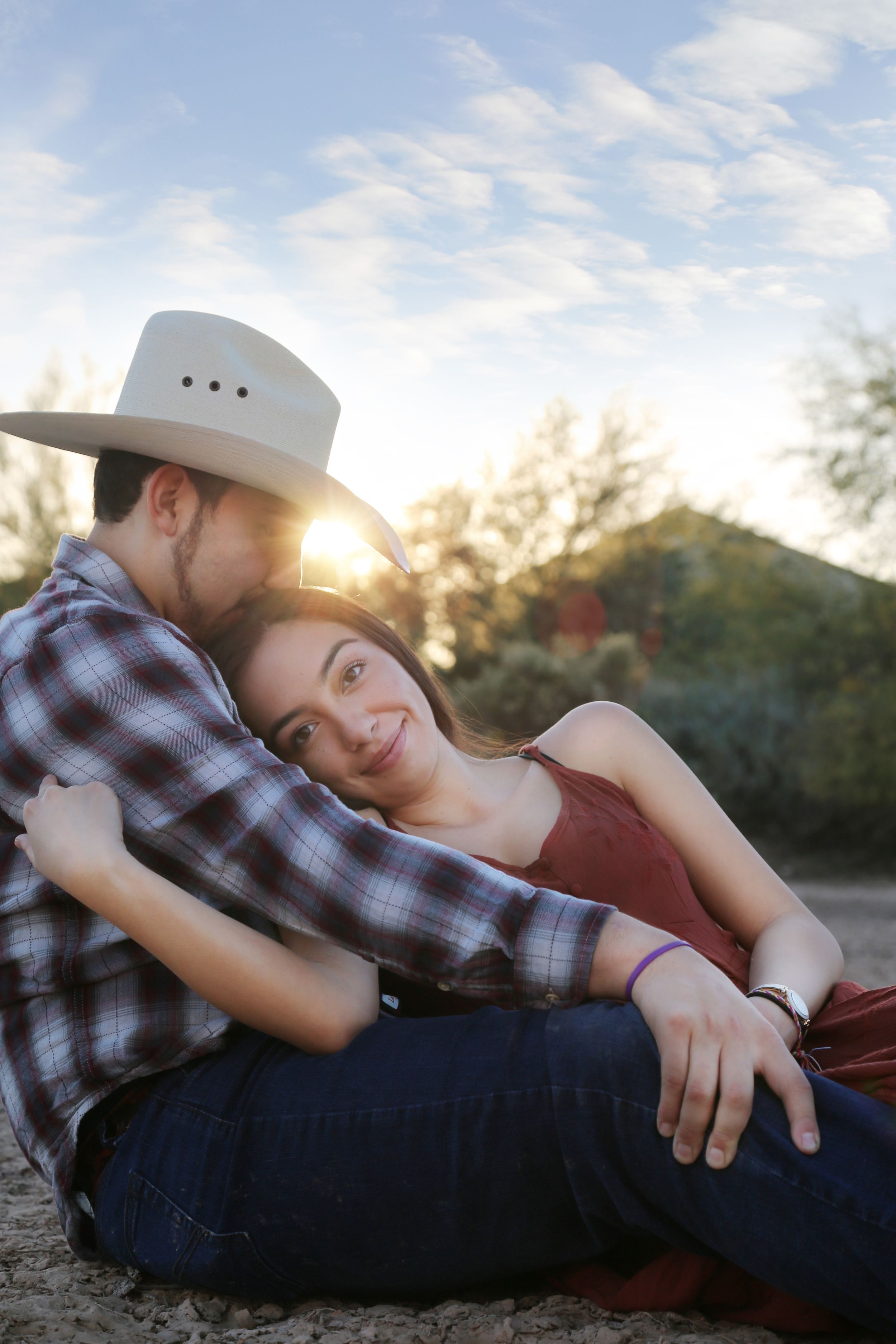 A smiling woman in a rust dress rests against a man in a cowboy hat and plaid shirt as the sun sets behind them.