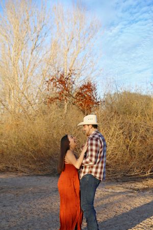 A couple in a cowboy hat, plaid shirt and flowing rust dress embrace on a dry desert path beside golden brush and bare trees.