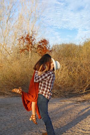 A man in a cowboy hat lifts his partner in a rust-colored dress into a kiss on a sandy trail with golden autumn brush behind them.