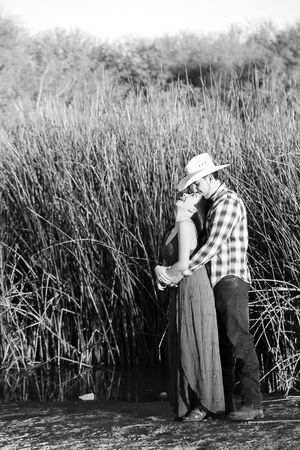 Black and white portrait of a couple embracing in front of tall marsh reeds, the man in a cowboy hat and plaid shirt.