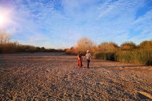A couple holds hands and walks across a vast dry riverbed at sunset under a wide blue sky with wispy clouds.