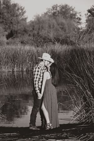 Black and white image of a couple kissing at the edge of a calm reed-lined pond, the man in a cowboy hat and plaid shirt.
