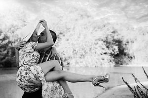 Black and white photo of a man lifting a woman in a floral dress as they kiss beside a lake, her hat held in her hand.