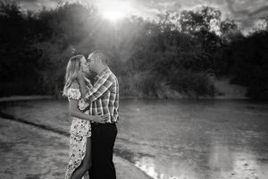 Black and white photo of a couple kissing by a shallow lake with bright sunbeams breaking through the trees behind them.