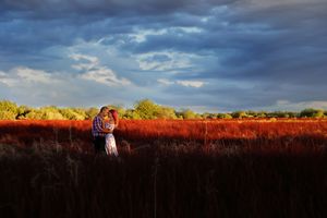 A couple embraces in a glowing red grass field under dramatic storm clouds, lit by warm golden sunlight.