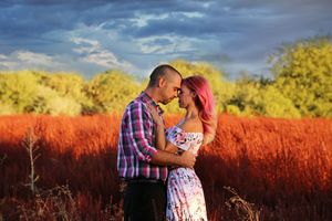 A man in a plaid shirt embraces a pink-haired woman in a floral dress in a vivid red field beneath a moody blue sky.