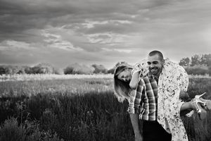 A smiling couple laughs and embraces in a grassy field under a moody sky in a black and white engagement photo.