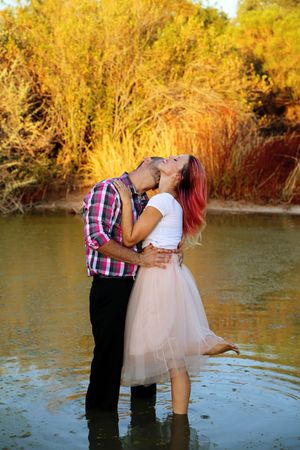 A couple in plaid and a tulle skirt kisses while standing in shallow water surrounded by golden autumn foliage.