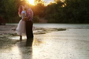 A couple embraces and kisses in shallow water at sunset as golden sunlight glows through the trees behind them.