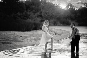 A woman in a white dress dips her foot in rippling water as a man watches in a black and white couples portrait.