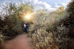 A couple embraces on a dirt path through desert shrubs as the sun sets, creating a warm glow in the background.