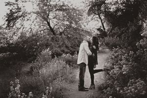 A couple kisses on a garden path lined with blooming bushes and trees in a vintage-style black and white portrait.