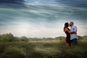 Couple embracing and kissing in a grassy field beneath a dramatic, swirling blue sky at sunset.