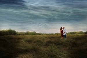 A couple kisses in a wide grassy meadow beneath a dramatic blue sky with wispy clouds in an engagement photo.