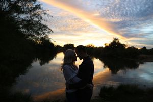 A silhouetted couple embraces beside a calm lake at sunset as colorful clouds reflect in the water.