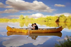 A couple sits together in a golden canoe on calm water, with trees and a bright blue sky reflected in the lake.
