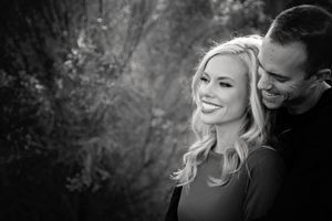 A smiling blonde woman laughs as her partner nuzzles close behind her in a black and white engagement portrait.