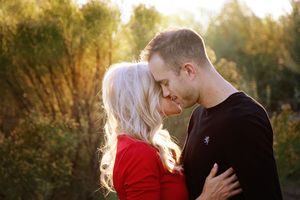 A man gently kisses the forehead of a blonde woman in a red top as warm sunlight glows through the trees behind them.