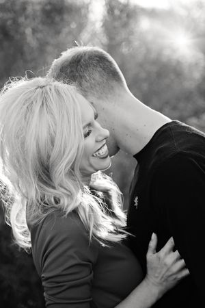 Black and white photo of a smiling blonde woman embraced by a man as sunlight streams through the trees behind them.