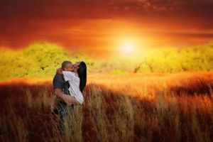 A couple embraces in a tall grass field beneath a glowing orange sunset sky, creating a romantic silhouette.