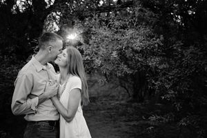 Black and white engagement photo of a couple embracing and nearly kissing under dark trees with sunlight peeking through.