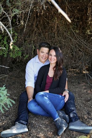 Happy couple sitting close together under leafy branches, smiling at the camera in a casual outdoor engagement portrait.