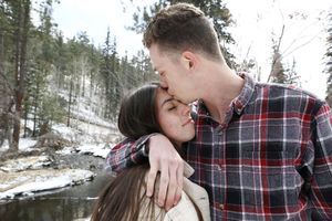 Man in a plaid flannel shirt kisses a woman's forehead in a snowy forest beside a stream during a winter engagement session.