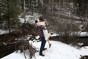 Man in plaid lifts and hugs a woman in a white dress on a snowy creek bank surrounded by evergreen trees.