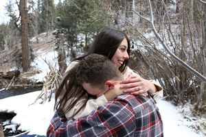 Laughing woman wearing an engagement ring hugs a man in a plaid shirt in a snowy forest setting.