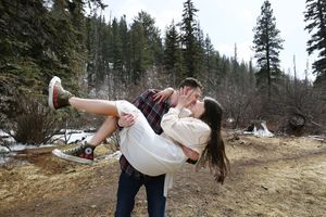 Man in plaid shirt and jeans cradles a woman in a white dress and sneakers as they kiss among tall pine trees.