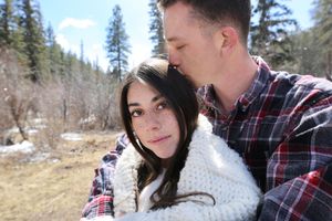 A man in a plaid shirt gently kisses the forehead of a smiling dark-haired woman in a cozy white knit sweater outdoors.
