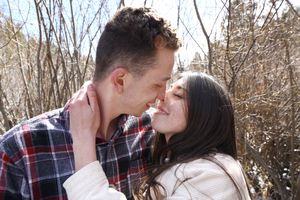 A couple in warm clothing share an intimate moment nose to nose among bare winter branches on a sunny day.