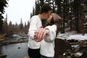A couple kisses beside a snowy creek in a pine forest while showing off the woman's sparkling engagement ring.