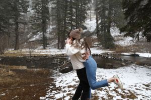 A man lifts his partner in a joyful embrace on a snowy forest trail beside a calm stream surrounded by evergreen trees.