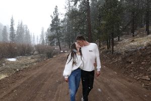 A couple kisses while walking along a dirt road through a misty pine forest with light snow falling around them.