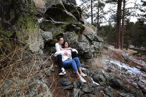 A couple sits close together on a mossy rock formation in a snowy forest, sharing a quiet moment during an engagement shoot.