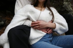 A close-up of a woman in a white knit sweater and jeans, her hands clasped with her partner's, showing an engagement ring.