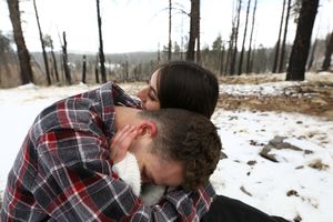 A couple in plaid and white embrace tenderly on snow-covered ground surrounded by bare trees in a quiet winter forest.