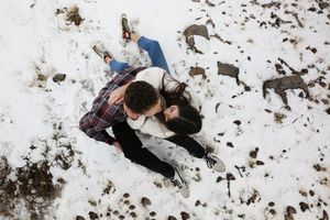 An overhead view of a couple lying close together on snowy ground, dressed in plaid and white sweaters, smiling at each other.