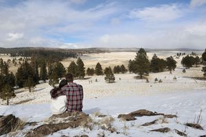 Couple sits close together on a snowy hillside rock, gazing across a wide open valley dotted with pine trees under a partly cloudy sky.