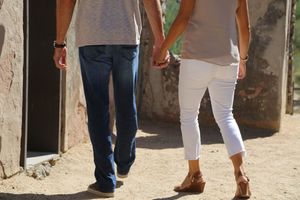 Couple holds hands while walking away along a sunlit dirt path beside old weathered stone walls, shown from the waist down.