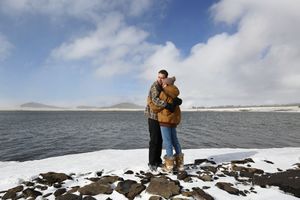 Couple in warm winter clothing hugs on a rocky, snow-dusted lakeshore with a calm lake and cloudy blue sky behind them.