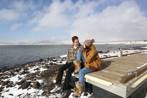 Couple sits together on a wooden dock beside a snowy lakeshore, smiling at each other beneath a blue sky with wispy clouds.